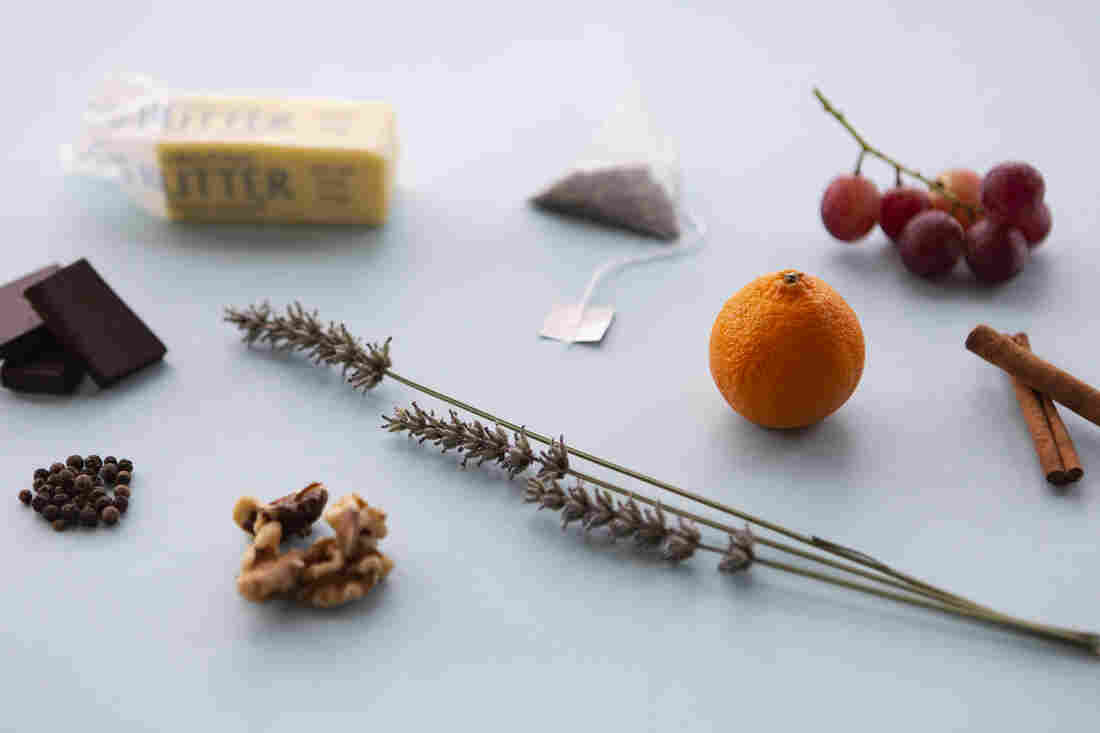 A tea bag, grapes, an orange, cinnamon sticks, lavender, walnuts, peppercorns, dark chocolate and butter are photographed at a 3/4 angle on a light blue background.