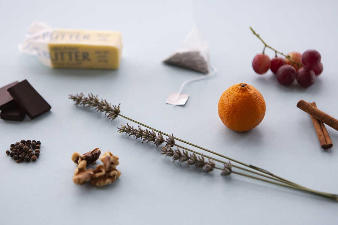 A tea bag, grapes, an orange, cinnamon sticks, lavender, walnuts, peppercorns, dark chocolate and butter are photographed at a 3/4 angle on a light blue background.