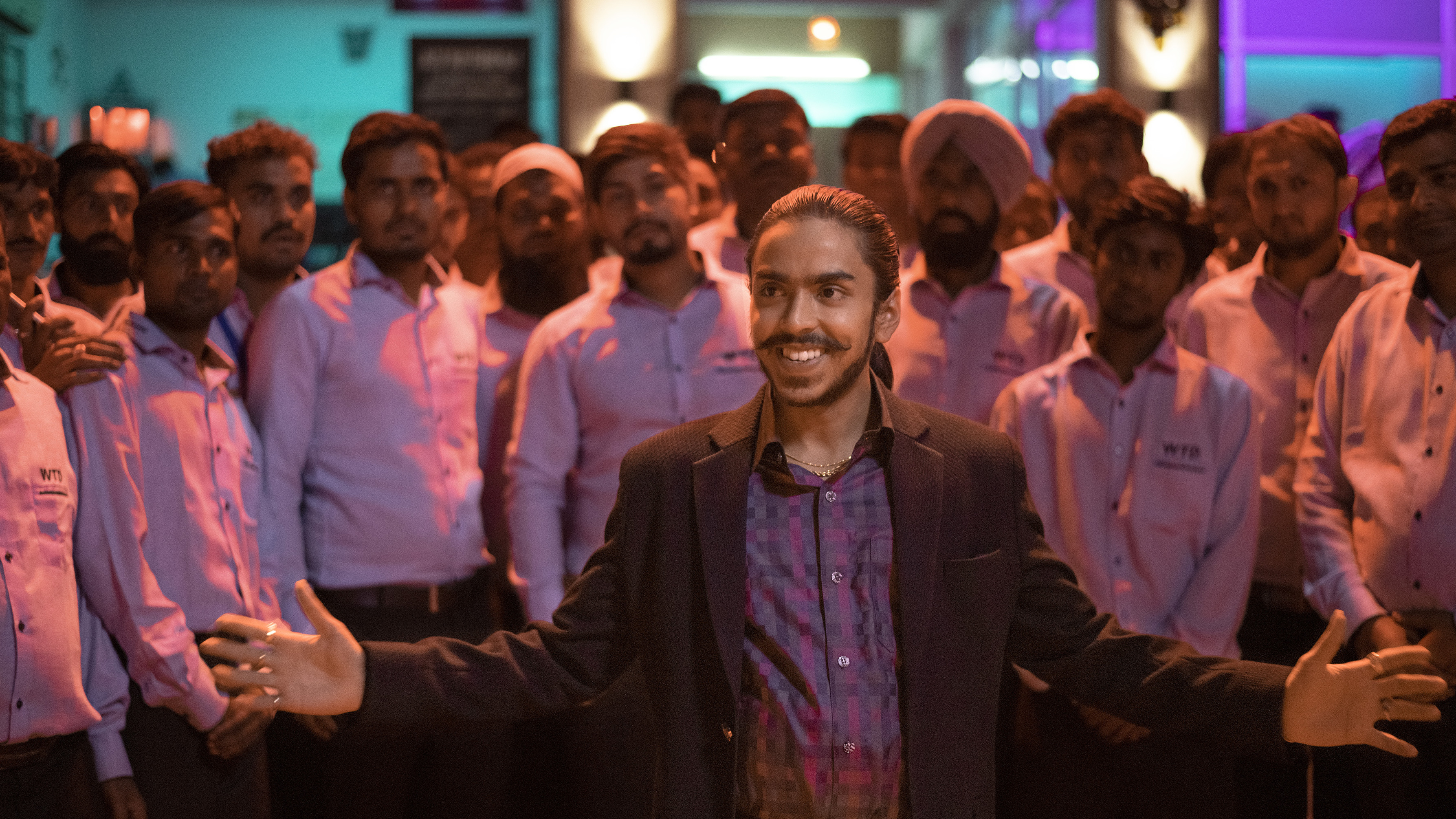 Balram smirks and opens his arms wide while standing in front of his taxi drivers and showing off his small empire.