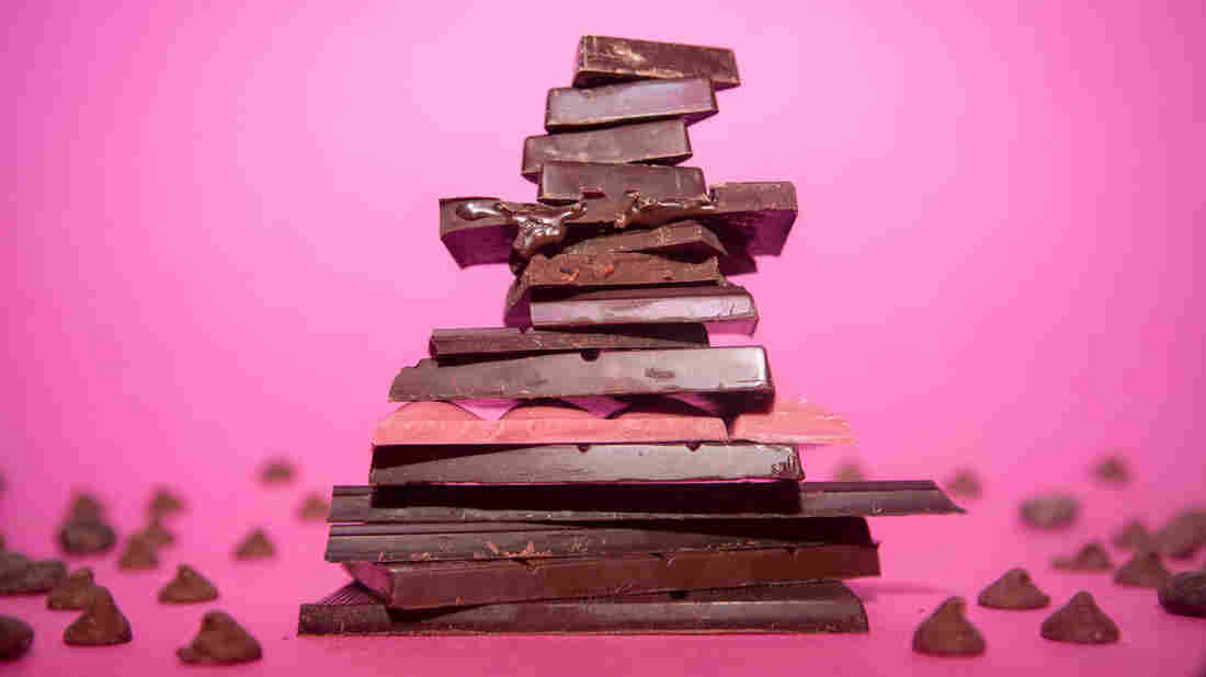 A tower made of stacked pieces of chocolate on a pink background with chocolate chips scattered around the base.