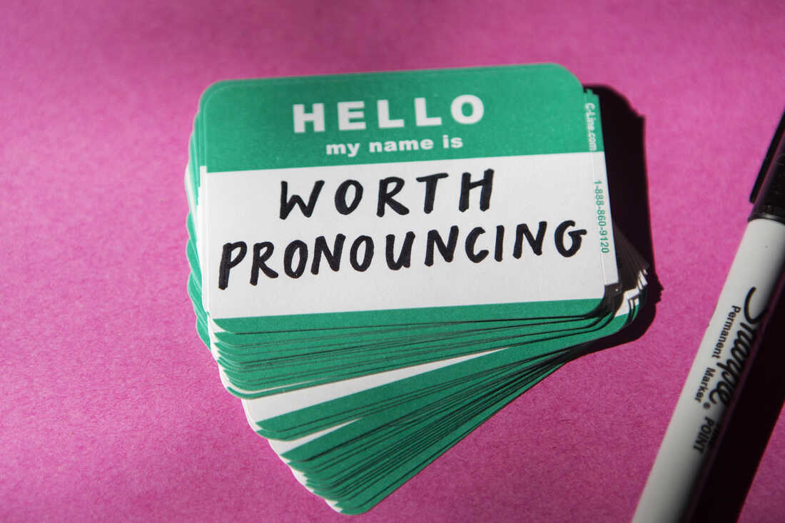 A teal name tag that reads "Hello my name is" is filled out "worth pronouncing." The name tag sits on a stack of other name tags against a magenta background.