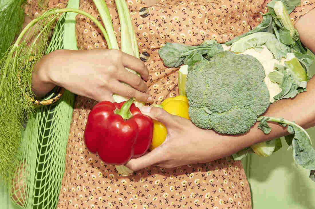 A woman wearing an orange floral, button-down dress carries a green mesh bag full of produce and holds extra produce in her arms.