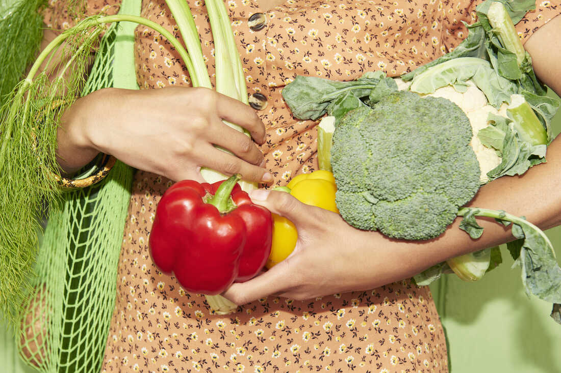 A woman wearing an orange floral, button-down dress carries a green mesh bag full of produce and holds extra produce in her arms.