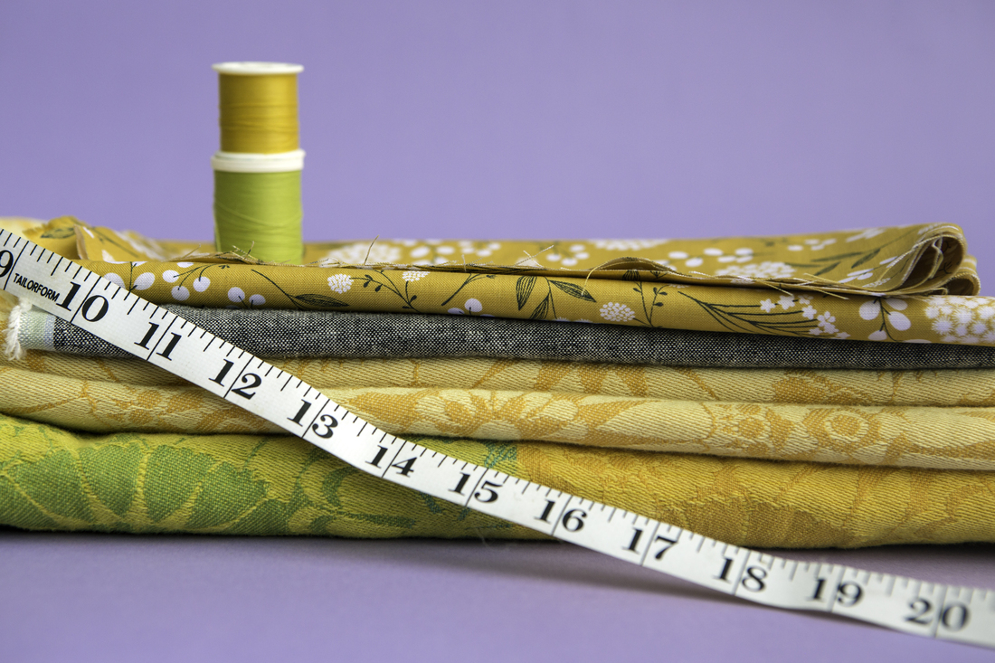 Three pieces of fabric in mustard yellow, dark gray, and bright yellow and green are stacked against a light purple backdrop. Two spools of thread in pea green and marigold yellow are stacked on top of the fabric, and a measuring tape is draped across the front.