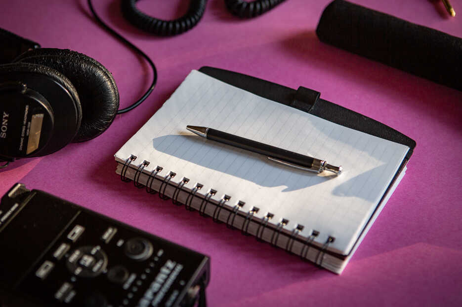 A blank notebook with a pen, a recorder, headphones and a mic are laid out on a bright fuchsia background.