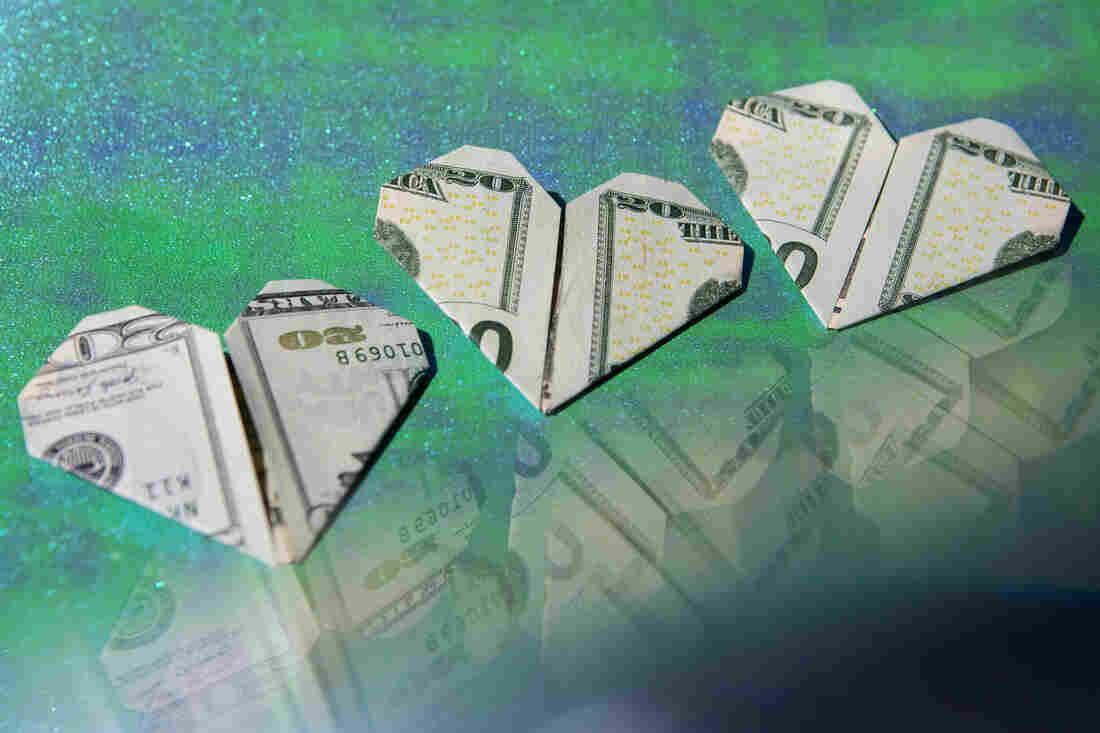 Three dollar bills folded into hearts are displayed on a sparkly green and blue background. A reflection of the money is seen in the bottom right corner of the frame.