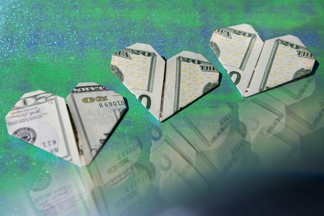 Three dollar bills folded into hearts are displayed on a sparkly green and blue background. A reflection of the money is seen in the bottom right corner of the frame.