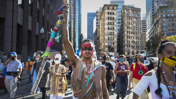 A man wearing face paint, a fringed vest and beaded necklaces lifts a ribbon-covered staff in the air while marching through city streets with other protesters. 