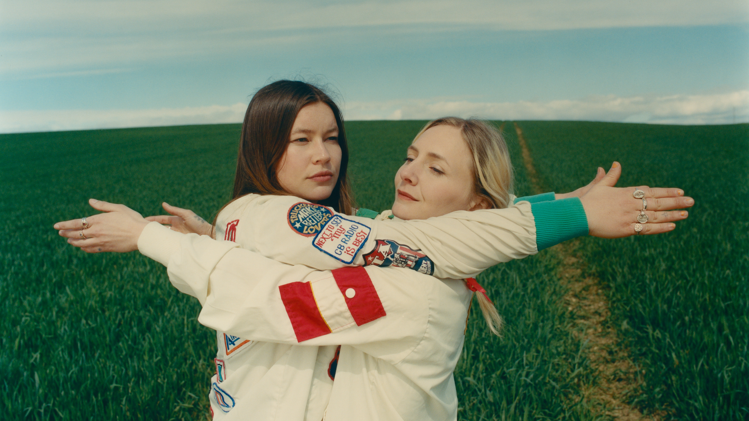 Rhian Teasdale (left) and Hester Chambers (right) embracing with their arms reaching past each other’s backs. They both are wearing long-sleeved shirts. The background shows grass and a clear blue sky.