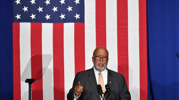 Rep. Bennie Thompson, wearing a suit, speaks into a microphone against a large vertical American flag.