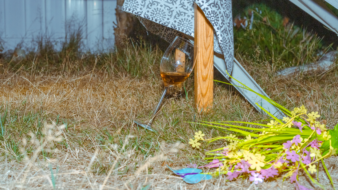 A half-glass of rosé tipped precariously on a picnic table leg. Flowers are strewn about the grass as if thrown in passion.