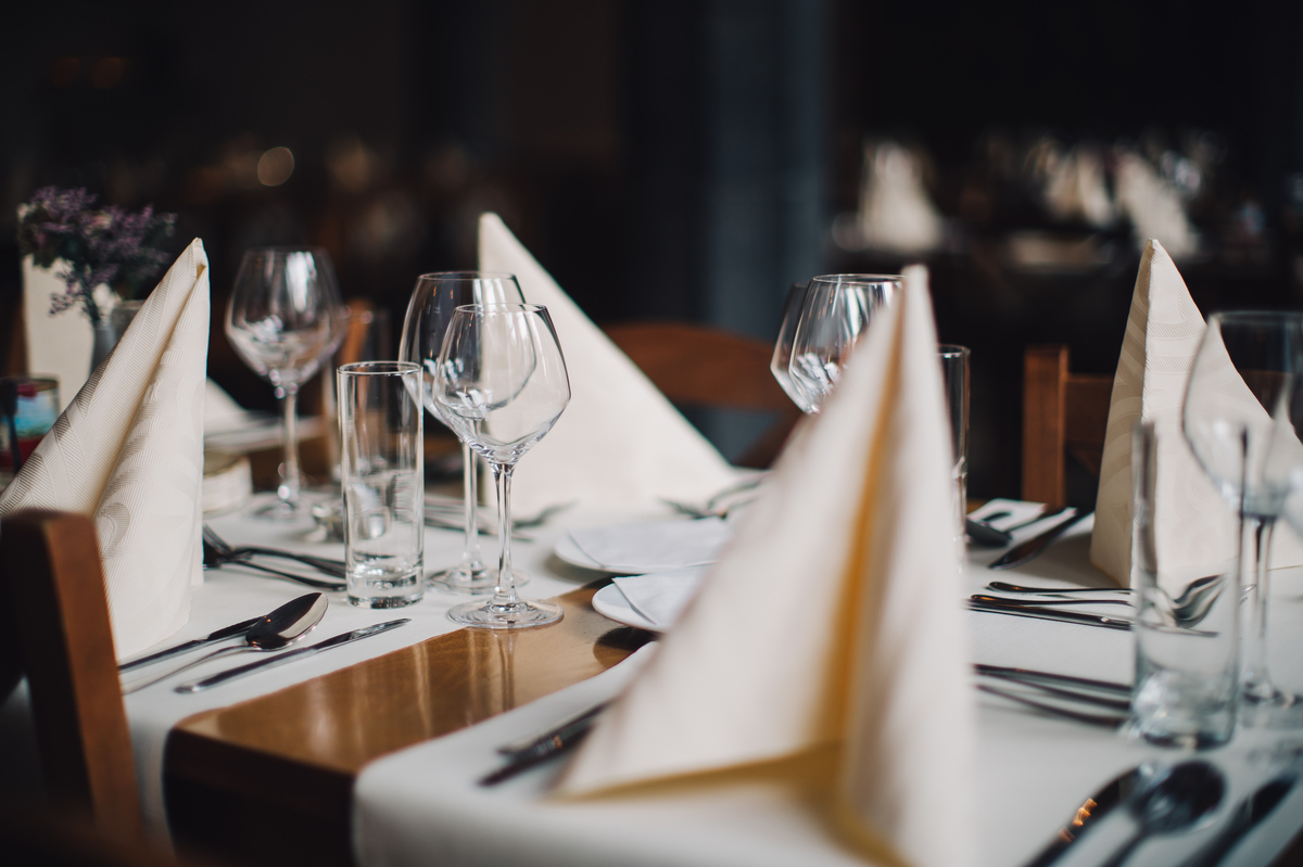 Des verres à vin et à eau brillants et des couverts sont sur la table en bois. Les serviettes en tissu sur les assiettes sont pliées en triangles. La table est recouverte d'une nappe.