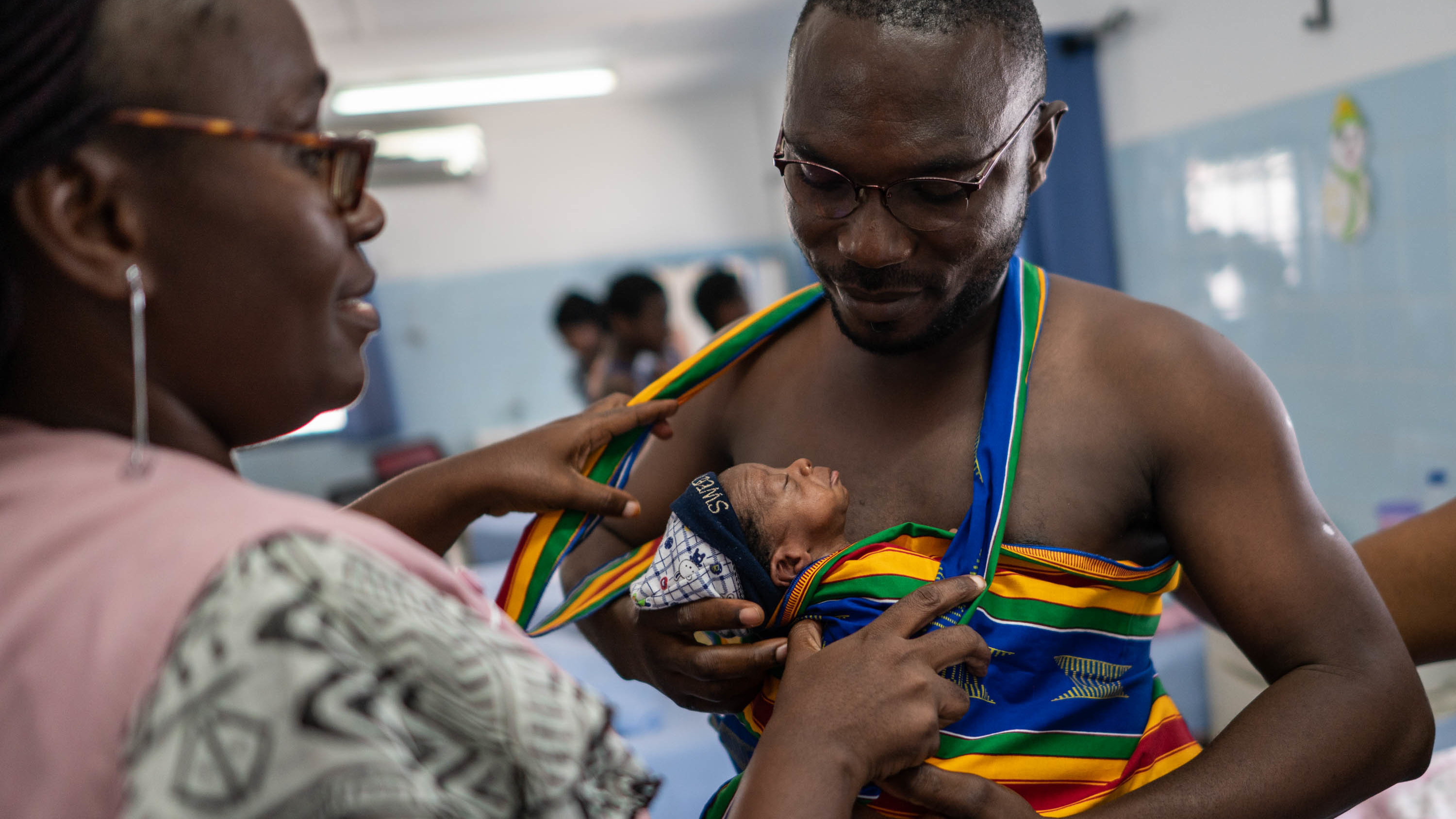 New father Yappe Pako gets help with his kangaroo care carrier from a midwife. His newborn son is named Ambo Crisostome. They