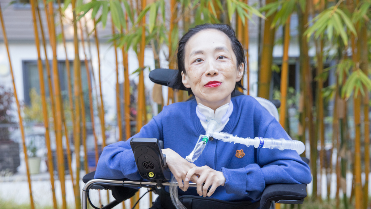 Photo of Alice Wong, an Asian American disabled woman in a power chair, against a background of bamboo trees. She is wearing a blue cardigan and sitting in a power chair. She is wearing a bold red lip color and a trach at her neck.