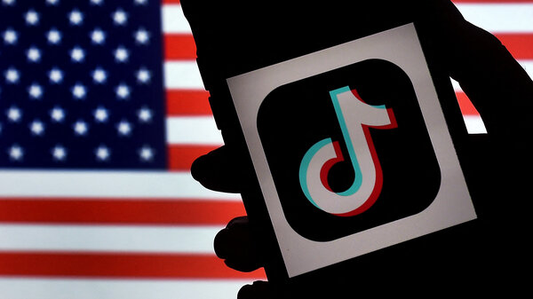 A silhouette of a hand holds a phone in front of an American flag. The phone screen shows the TikTok logo on a white background. 