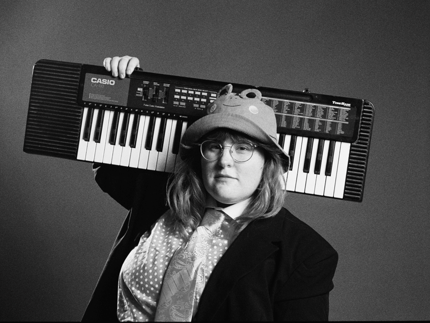 Le musicien corook porte une chemise, une veste et une cravate, ainsi que son chapeau seau grenouille signature. Ils tiennent un clavier sur leur épaule.