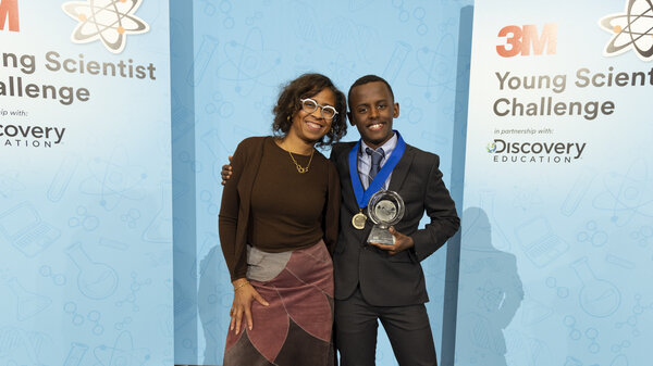 Heman Bekele, a young man in a black suit, holds a trophy and smiles next to his 3M mentor, Deborah Isabelle (left), a woman wearing a brown shirt and skirt.