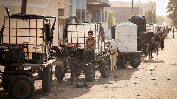 A boy in Khan Younis sits on a donkey-drawn cart loaded with a water tank. 