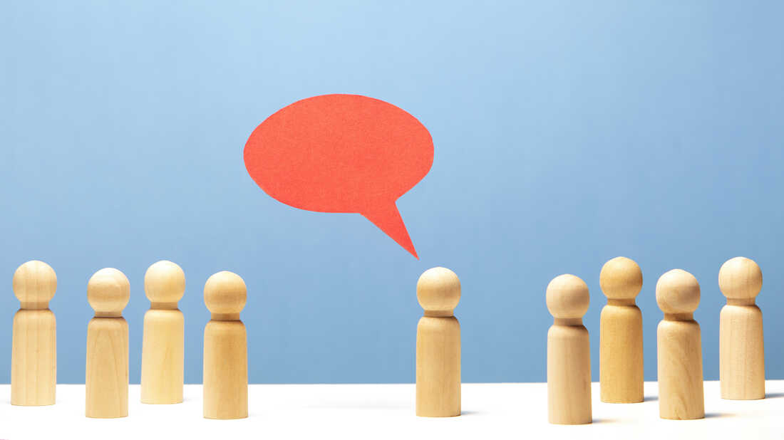 Red speech bubble in mid air over wooden figurine surrounded by 8 other figurines on white surface, blue background