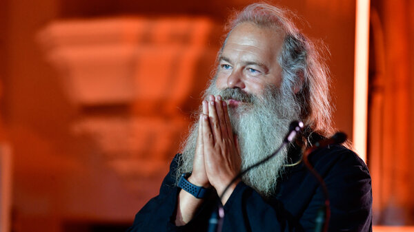 Rick Rubin, a man with a long beard, stands in front of a microphone. He holds his hands up to his face in a prayer position. 