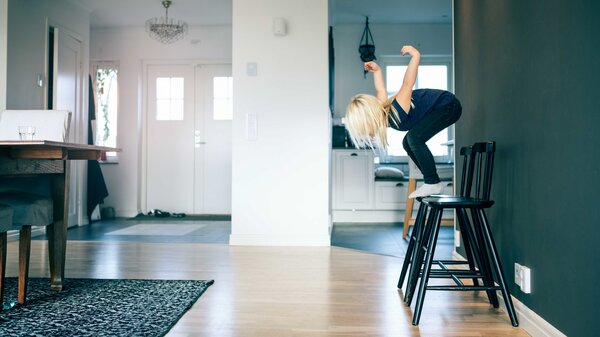 A child with long blond hair prepares to jump off of a chair.  