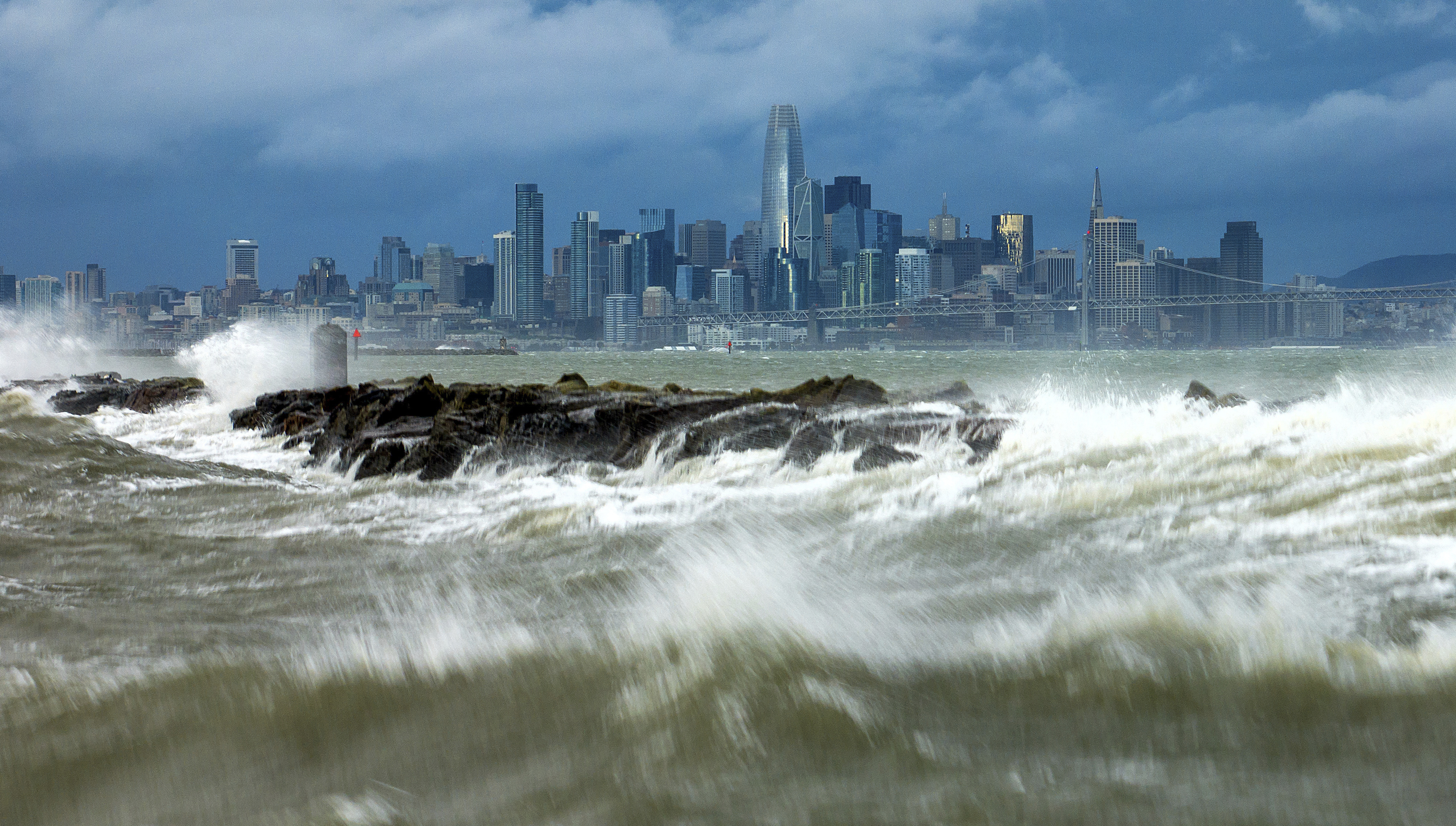 Alameda: Waves crash over a breakwater with the San Francisco skyline in the background on Sunday. High winds and heavy rainfall are impacting the region.