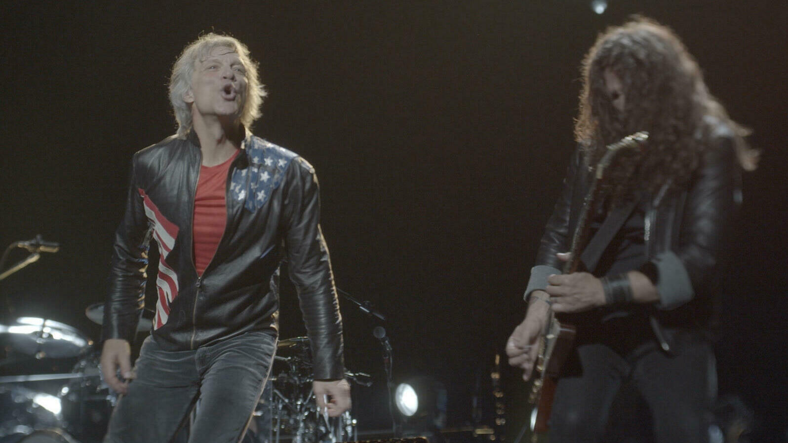A man with shaggy grey hair, wearing a leather jacket sings towards his audience. At his right is a long haired man playing the guitar.