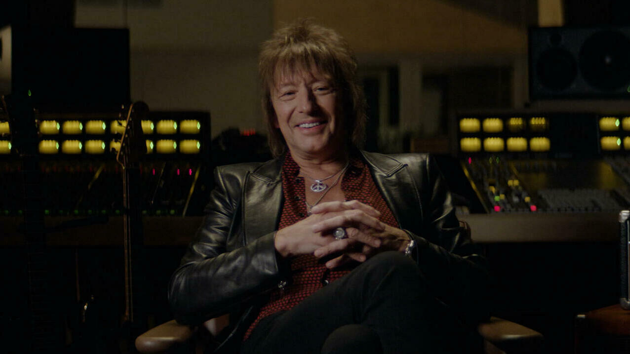 An older man sits in a recording studio smiling. He is wearing a leather jacket and unbuttoned red shirt.