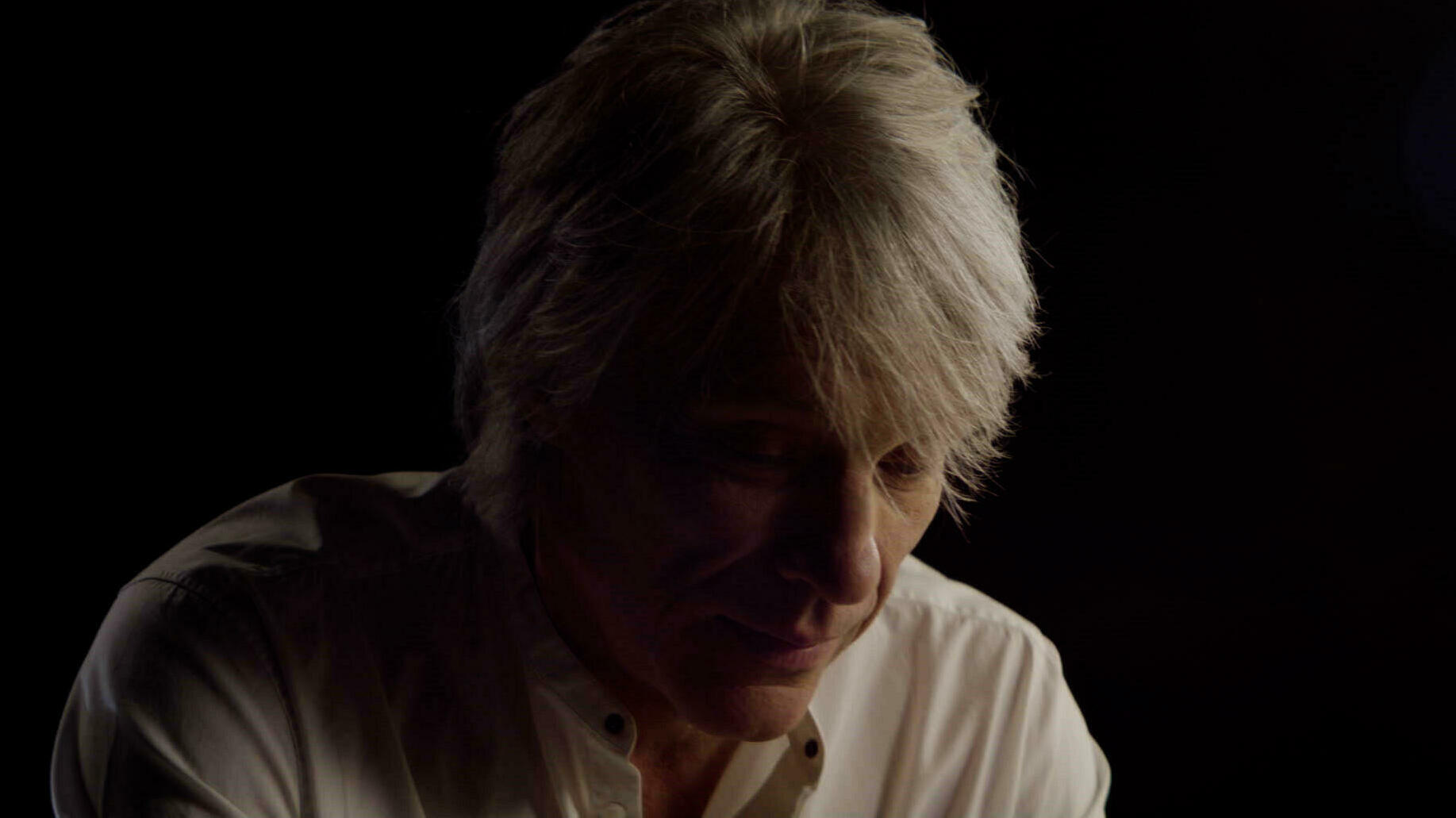 A man with shaggy grey hair sits pensively in a dark room, head bowed forward.