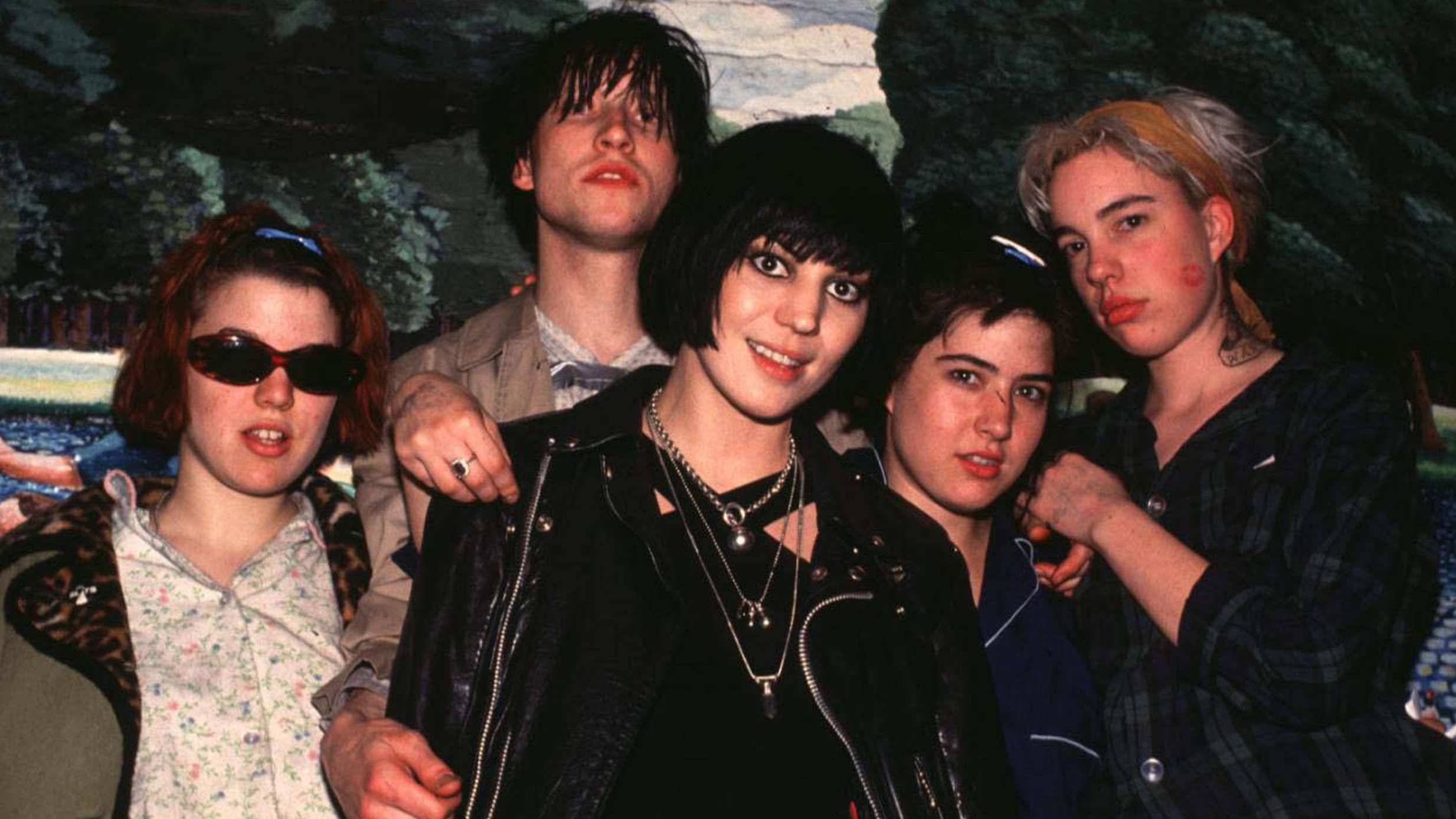 A girl with a black bob hair cut wearing a leather jacket stands surrounded by three young women and one young man.