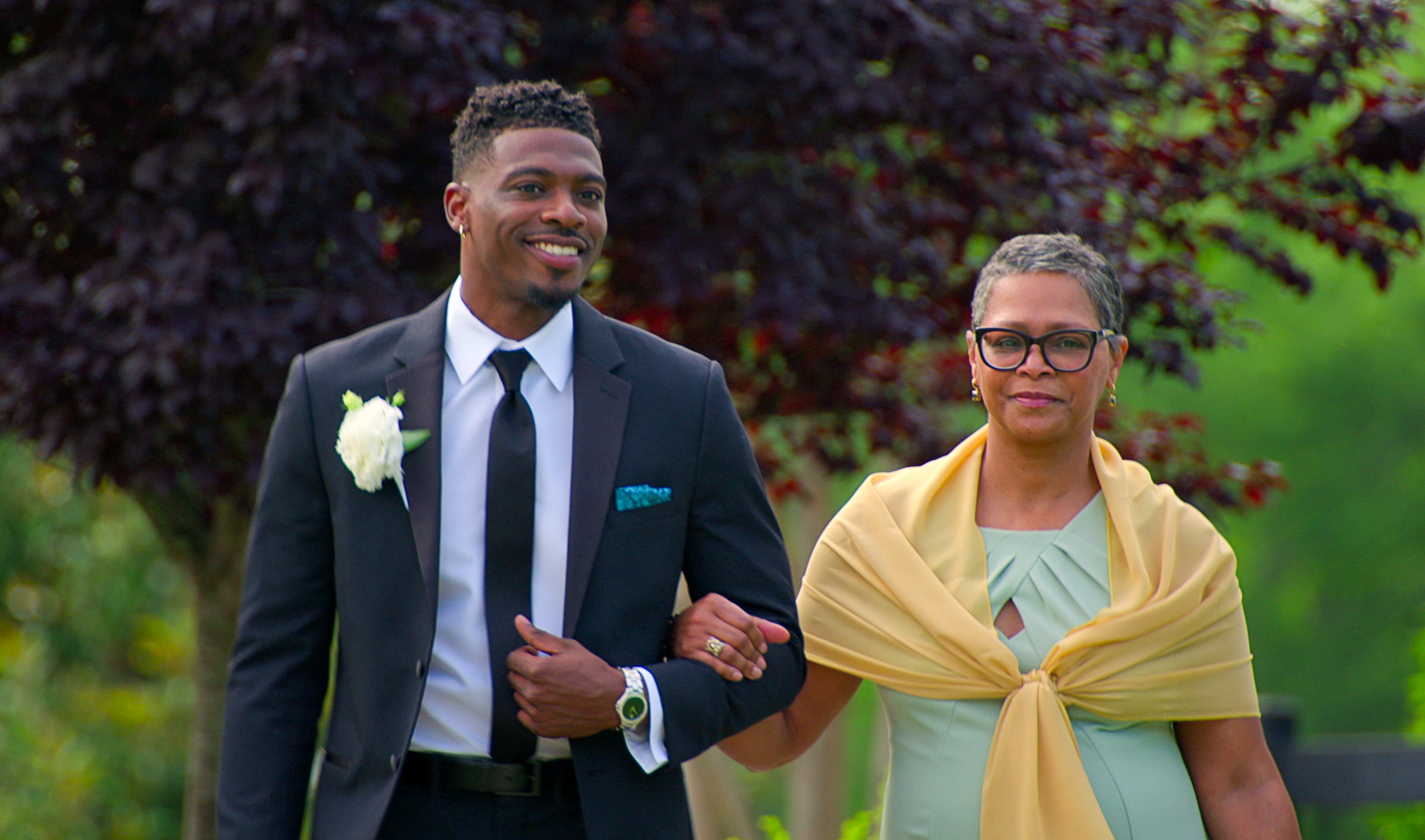 A young Black man in a smart suit walks arm-in-arm with an older Black woman who is wearing a green formal dress and yellow shoulder wrap.
