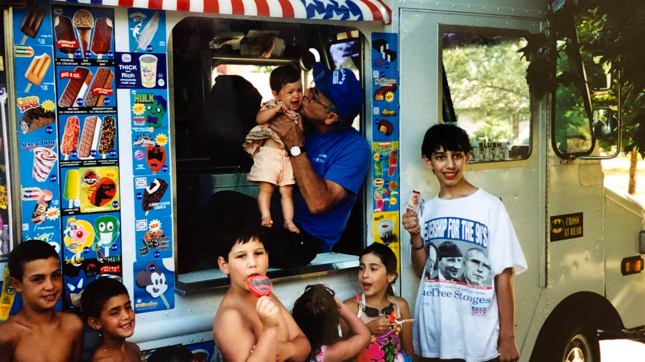 A man in a blue shirt and cap holds a baby in an ice cream truck and kisses them on the cheek. Outside of the truck are seven children, some of whom are holding and eating cold treats. 