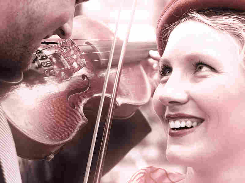 A young, smiling woman gazes up at a man playing violin