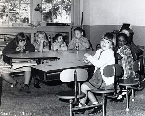 Seven children sit at an arced table, four facing camera and the other three turned around to look at camera.  Three of the children have Down Syndrome.