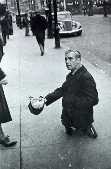 A middle-aged legless man "stands" on a sidewalk holding his hat out to passerbys..