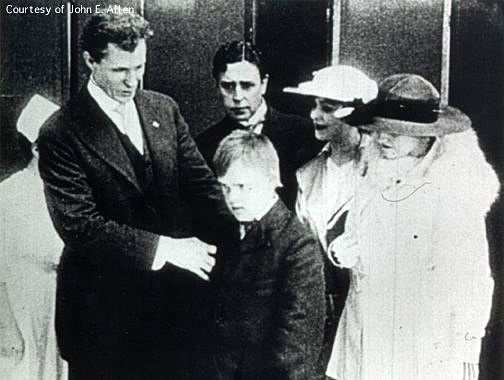 Group shot including: Dr. Haiselden, a nurse, a gentleman, two women, and a young boy with Down Syndrome, all in fine dress.  Dr. Haiselden  appears to be examining the boy; the women look on with pleasant expressions while the man wears a look of worry.